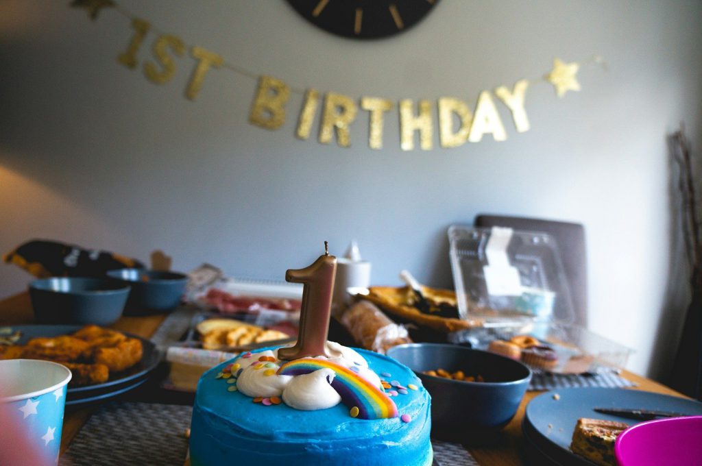 Bánh kem màu xanh dương cắm cây nến số 1 màu vàng đặt trên bàn tiệc, phía sau treo băng rôn 1st birthday