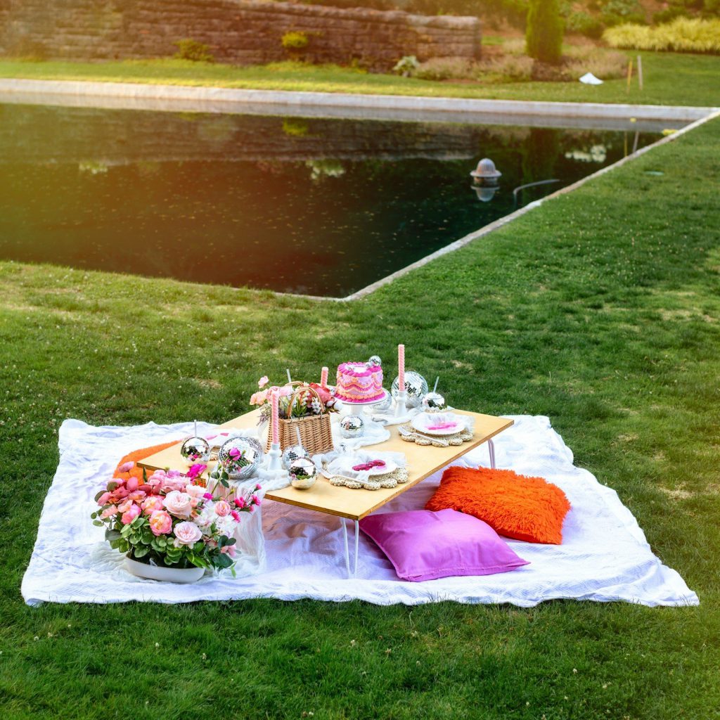 Bàn picnic thấp đặt trên tấm khăn trắng giữa bãi cỏ, có hoa và bánh kem